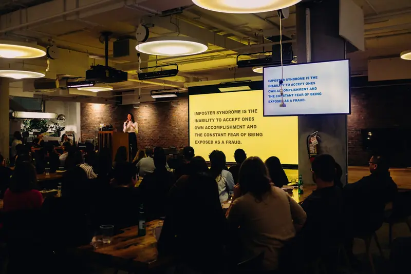 A woman speaking into a microphone to a seated audience, with slides about imposter syndrome on the screens