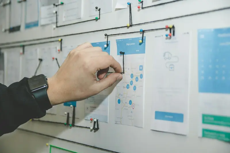 A hand stretching a string between pinned app screen sketches on a wall, linking them like a flow diagram