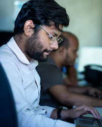 A man in glasses typing on a laptop in an office, with a colleague working behind him