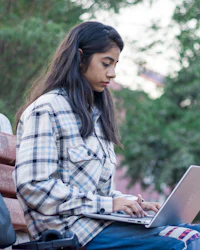 A young woman in a checked shirt typing on a laptop while sitting on a park bench