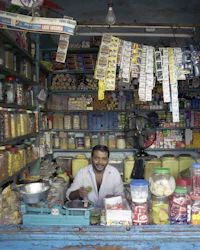 A smiling man behind the counter of a small general store packed with jars and hanging packets